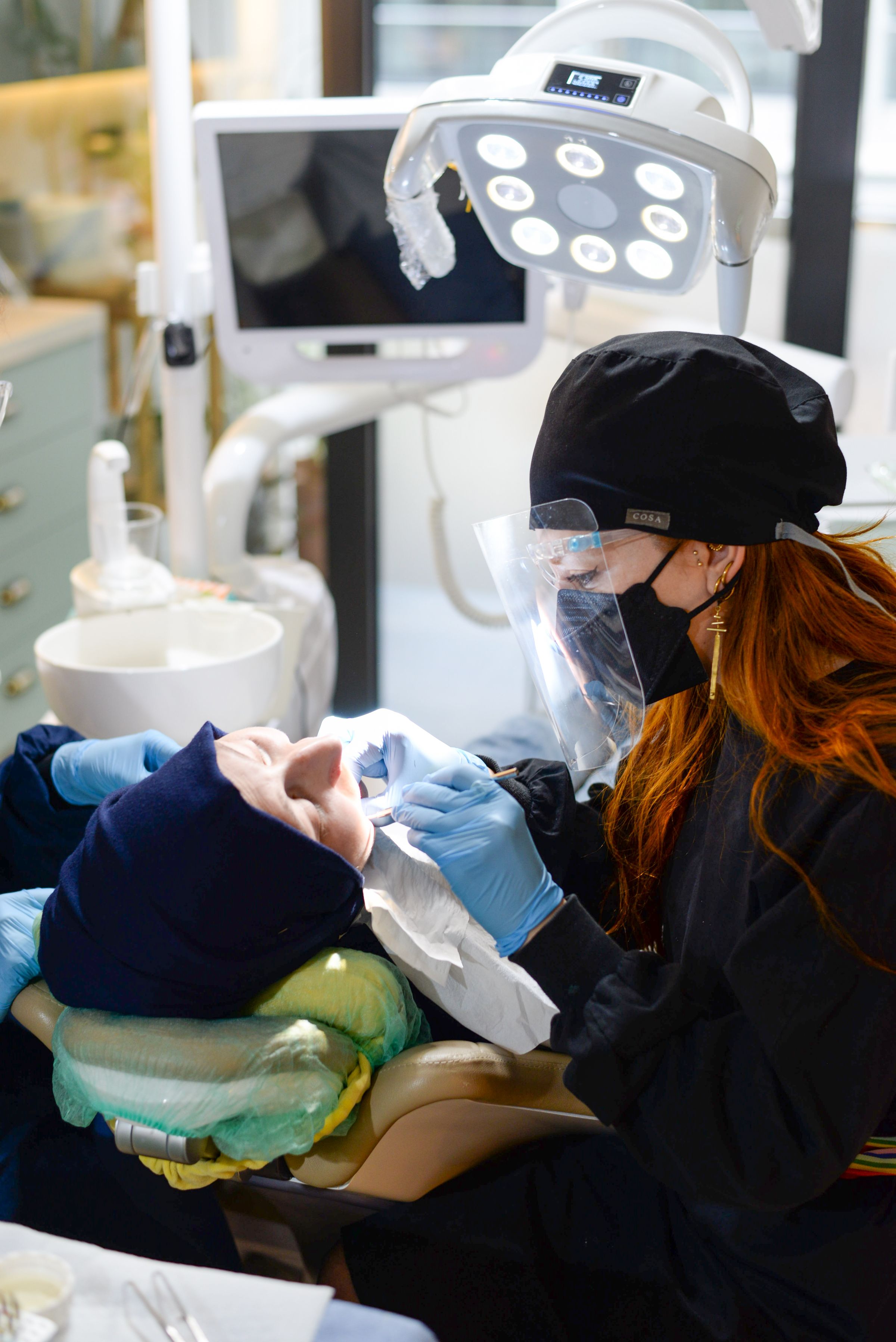 A dentist in a face shield examines a patient's teeth under an exam light with a chairside monitor in view.